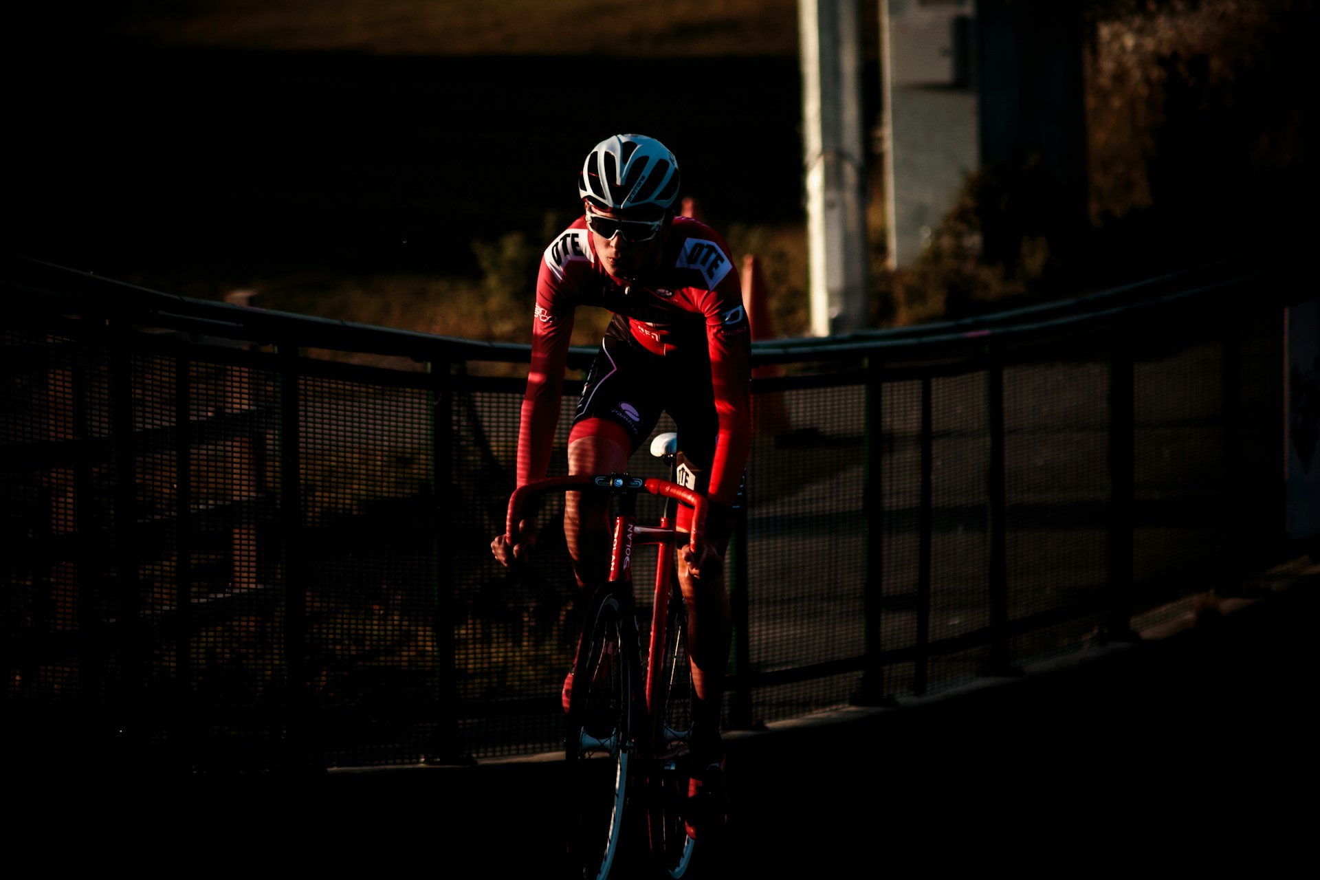 A cyclist in a red race kit riding through a narrow band of low evening light, with the track and its fencing falling away into darkness behind