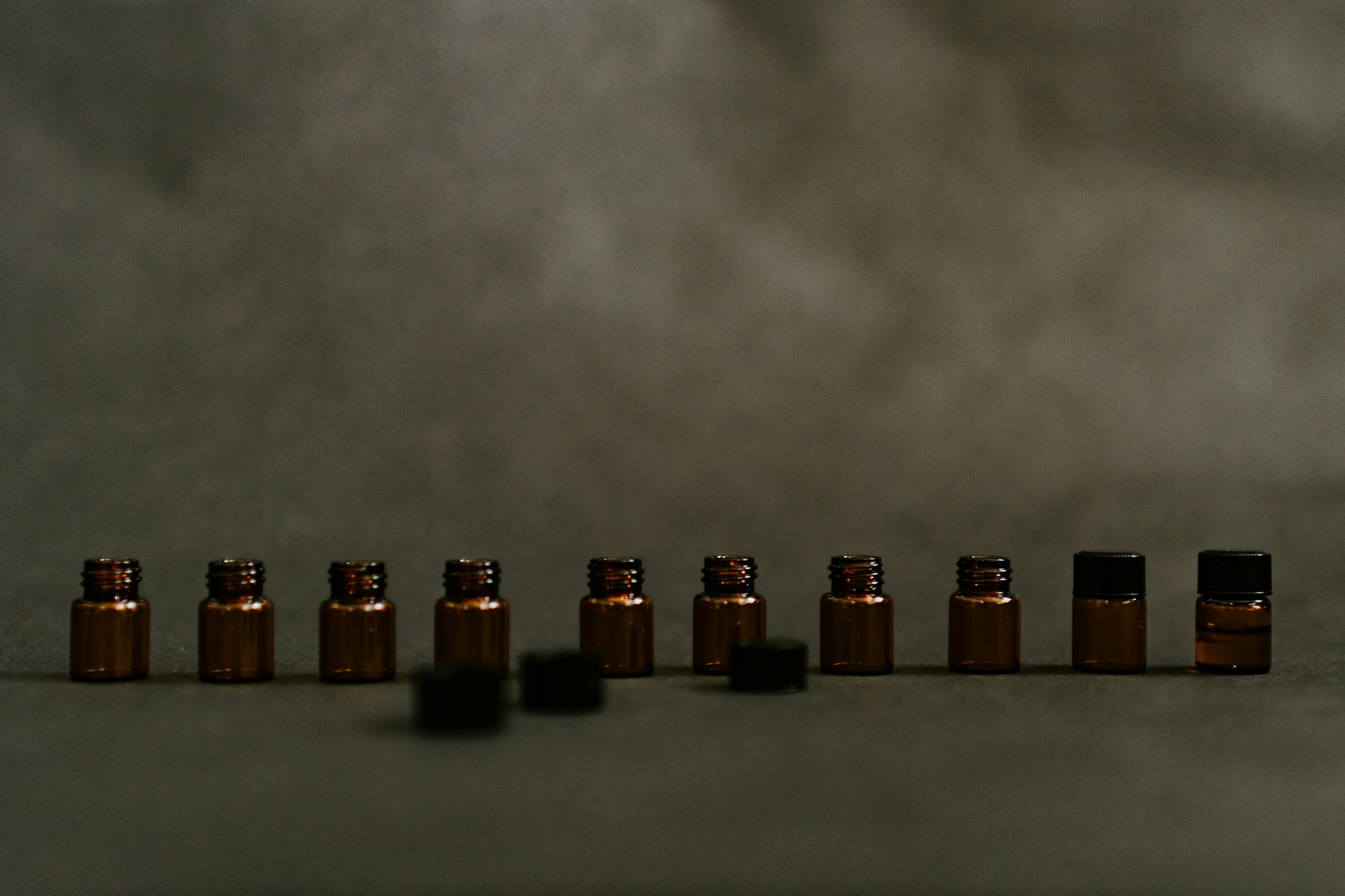 A row of small amber glass vials on a grey surface, most open, two capped, with three loose black caps lying in front of them